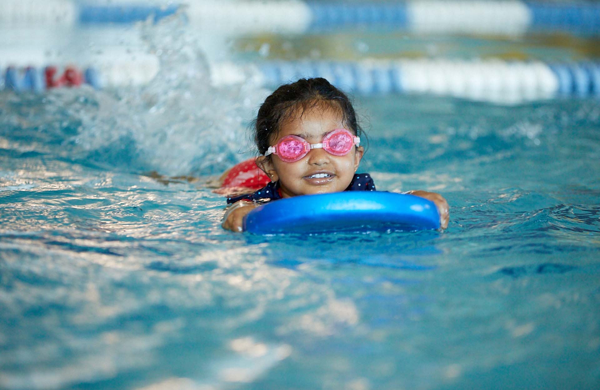 young Indian child wearing goggles using swimming kickboard in pool during swim lesson young Indian child wearing goggles using swimming kickboard in pool during swim lesson